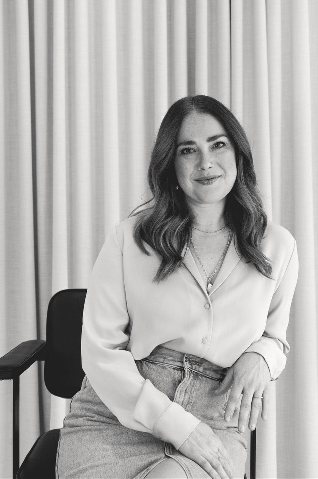 Black and white photo of a woman sitting in a chair against a curtain backdrop