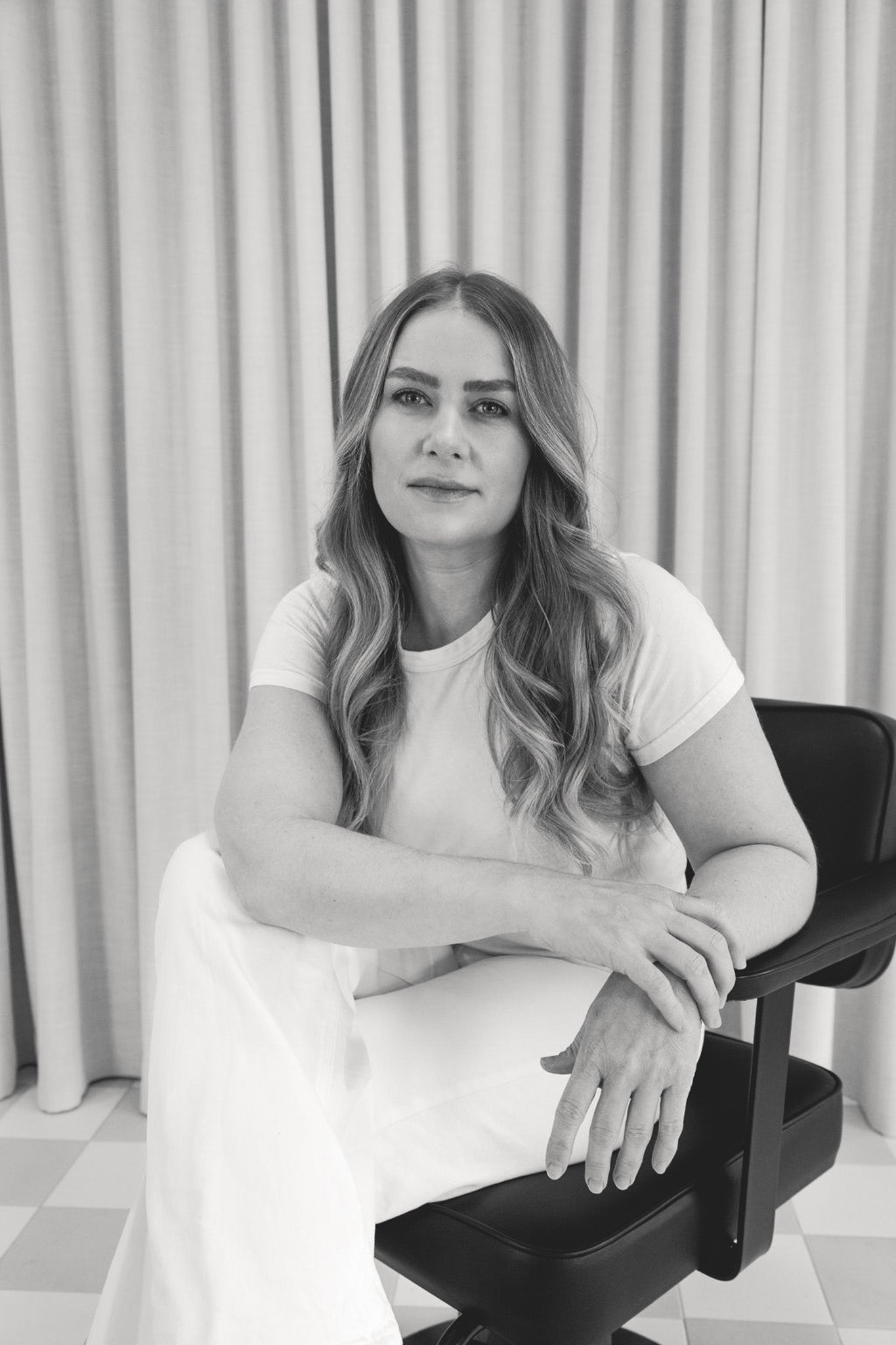 Black and white photo of a woman sitting on a chair against a curtain background