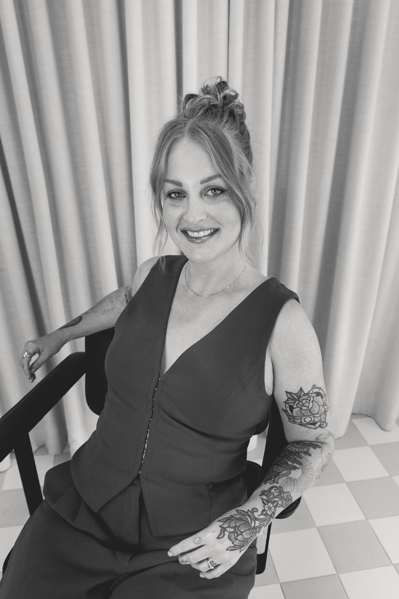 Black and white photo of a woman with tattoos sitting in a chair against a curtain backdrop.