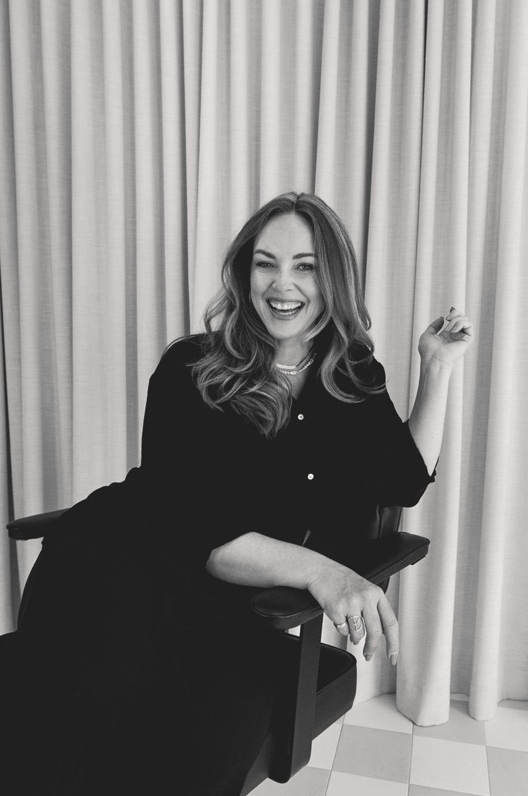 Black and white photo of a woman sitting on a stool in front of a curtain.