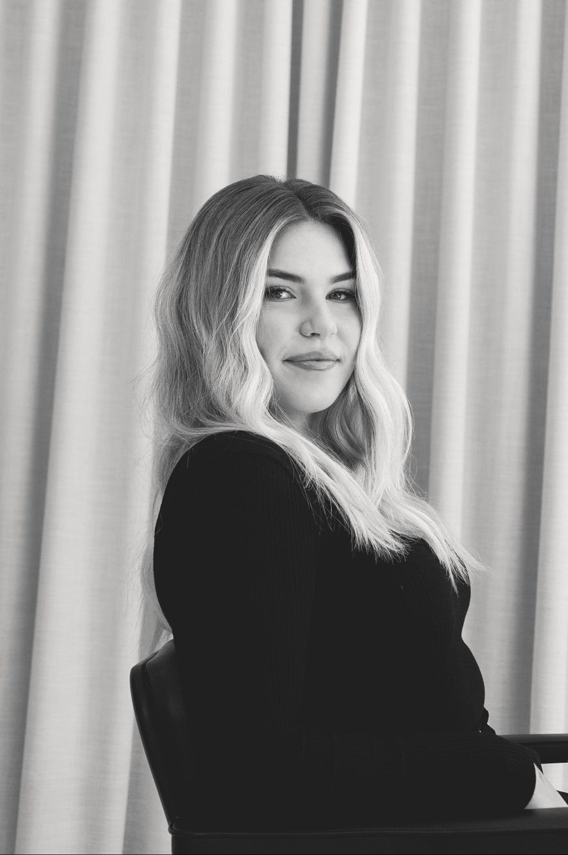 Black and white portrait of a woman sitting in front of a curtain.