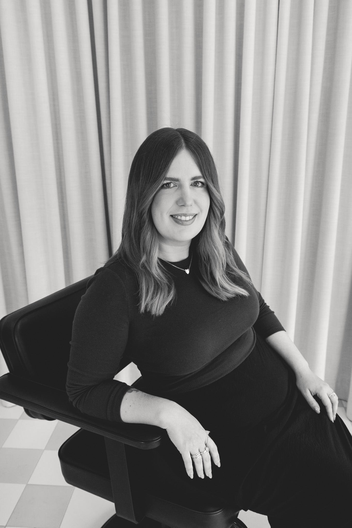 Black and white photo of a woman sitting on a chair with curtains in the background