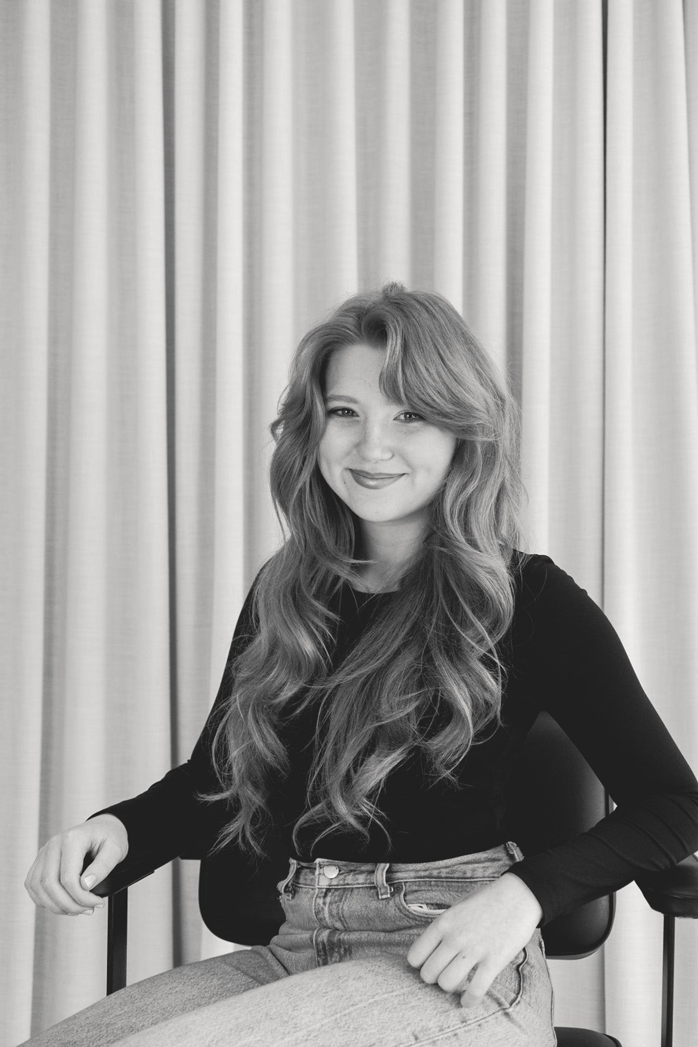 Black and white photo of a girl sitting on a chair with curtains in the background