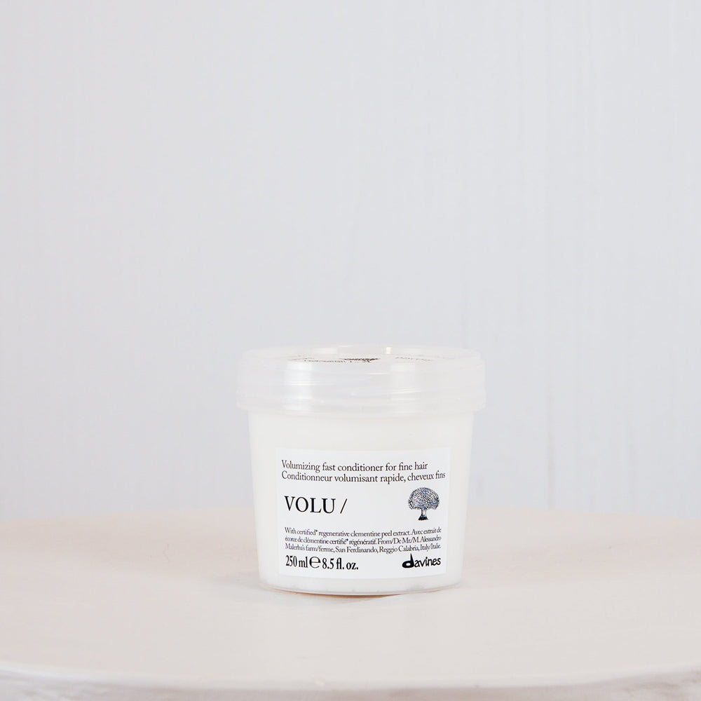 A small white cosmetic jar with a clear lid sits centered on a round white pedestal against a light gray textured background. The label shows the brand name VOLU.