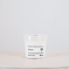 A small white cosmetic jar with a clear lid sits centered on a round white pedestal against a light gray textured background. The label shows the brand name VOLU.