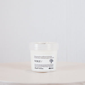 A small white cosmetic jar with a clear lid sits centered on a round white pedestal against a light gray textured background. The label shows the brand name VOLU.